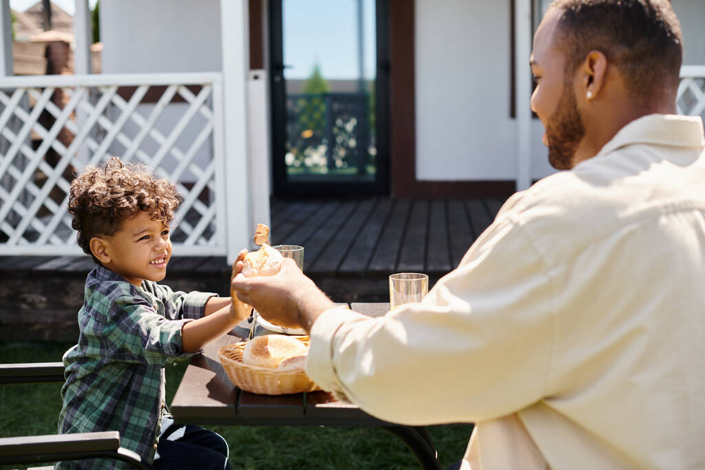happy african american father passing fresh bun to curly smiling son while having family bbq - Photo, Image