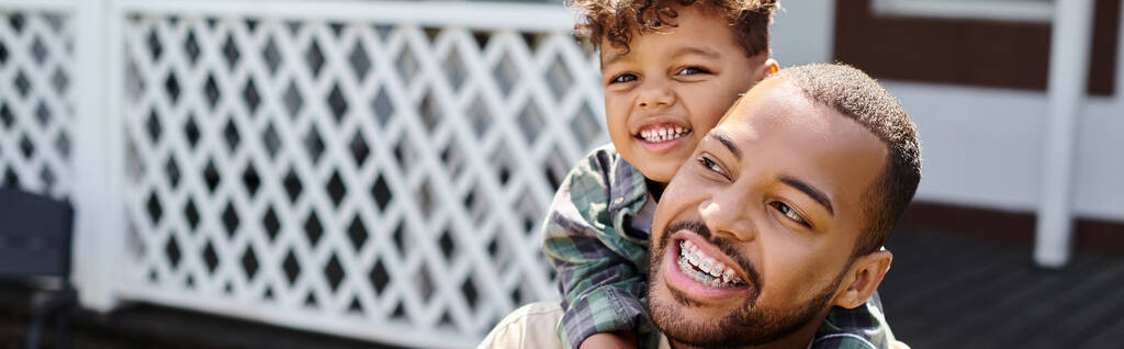 positivo afroamericano niño abrazando excitado padre en aparatos ortopédicos en el patio trasero de la casa, bandera - Foto, Imagen