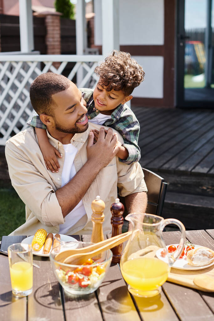 niño afroamericano positivo abrazando al padre excitado en aparatos ortopédicos en el patio trasero de la casa, tiempo en familia - Foto, Imagen