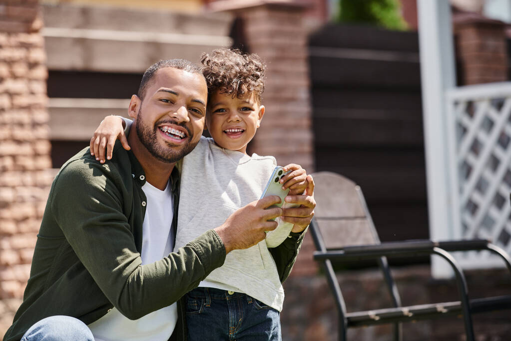 positivo uomo afroamericano in apparecchio acustico e figlio utilizzando smartphone sul cortile di casa in periferia - Foto, immagini