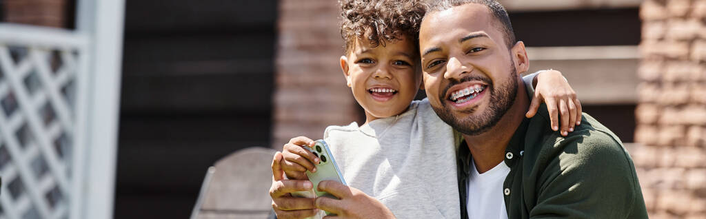 pozytywny Afrykański Amerykanin w aparacie na zęby i syn za pomocą smartfona na podwórku domu, baner - Zdjęcie, obraz