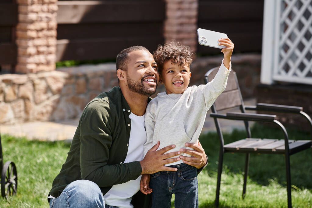 niño afroamericano feliz tomando selfie en teléfono inteligente con su padre en aparatos ortopédicos en el patio trasero - Foto, imagen