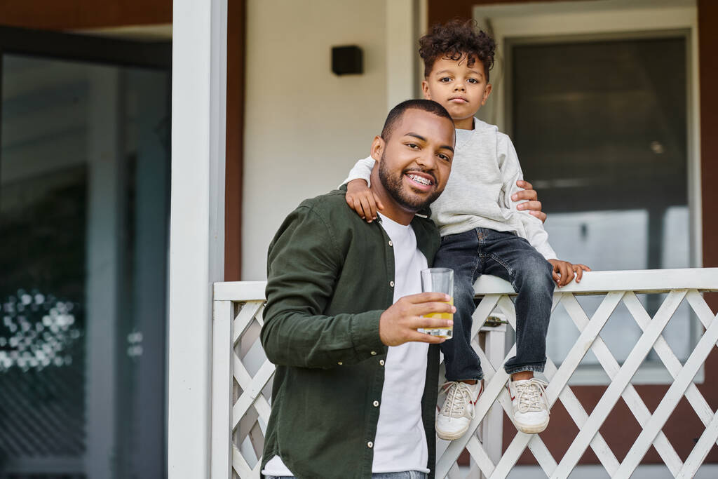 joyeux père afro-américain dans des bretelles tenant le jus d'orange et étreignant son fils assis sur le porche - Photo, image