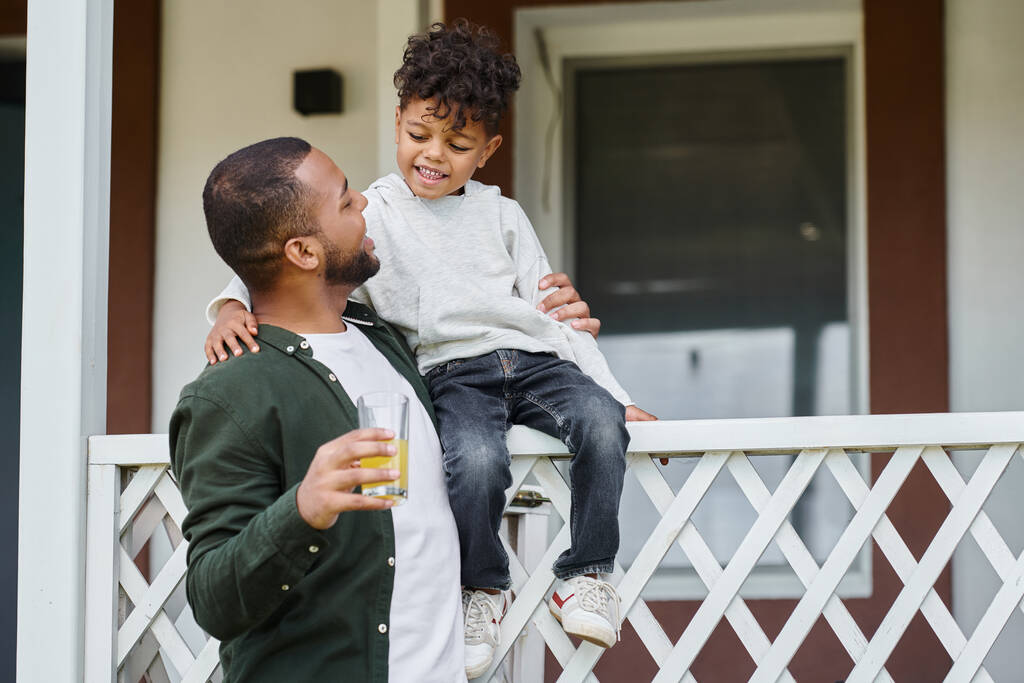 allegro padre afroamericano in bretelle che tiene il succo d'arancia e abbraccia suo figlio seduto sul portico - Foto, immagini