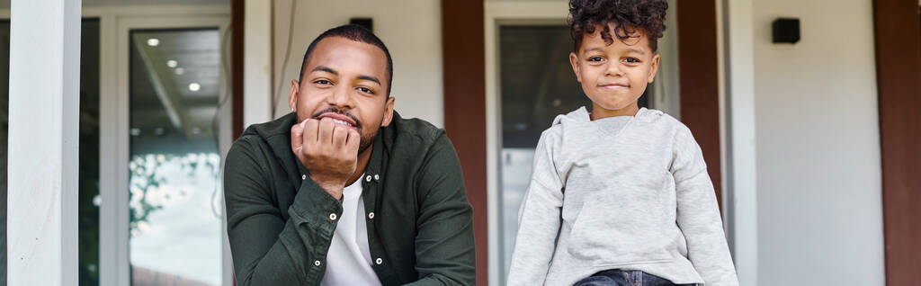 alegre afroamericano padre e hijo sonriendo y sentado en el porche de la casa, bandera de la familia - Foto, imagen