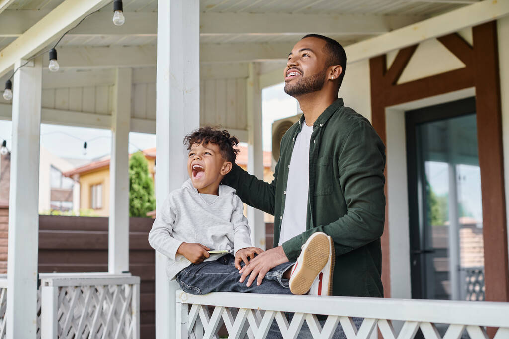 Heyecanlı Afrikalı Amerikalı baba ve oğul gülümsüyor ve verandada oturuyor, aile portresi - Fotoğraf, Görsel