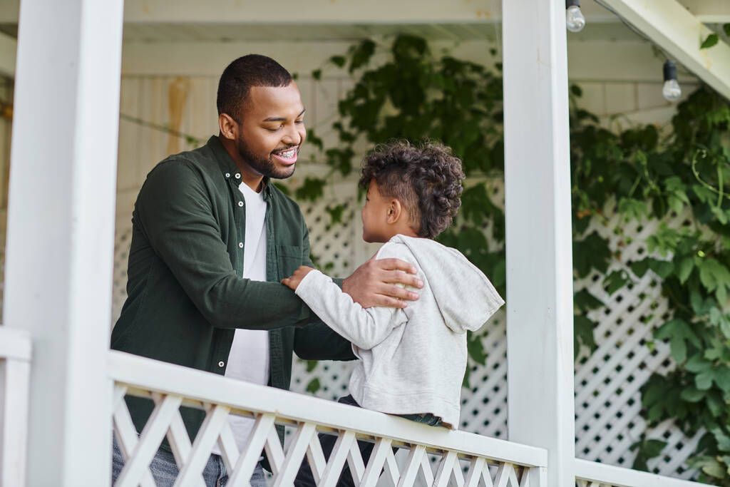 heureux père afro-américain dans des bretelles étreignant mignon fils bouclé assis sur le porche sur la maison dans les banlieues - Photo, image