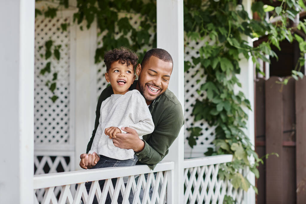 щасливий афроамериканський чоловік в брекетах сміється і обіймає кучерявого сина, сидячи на ганку - Фото, зображення
