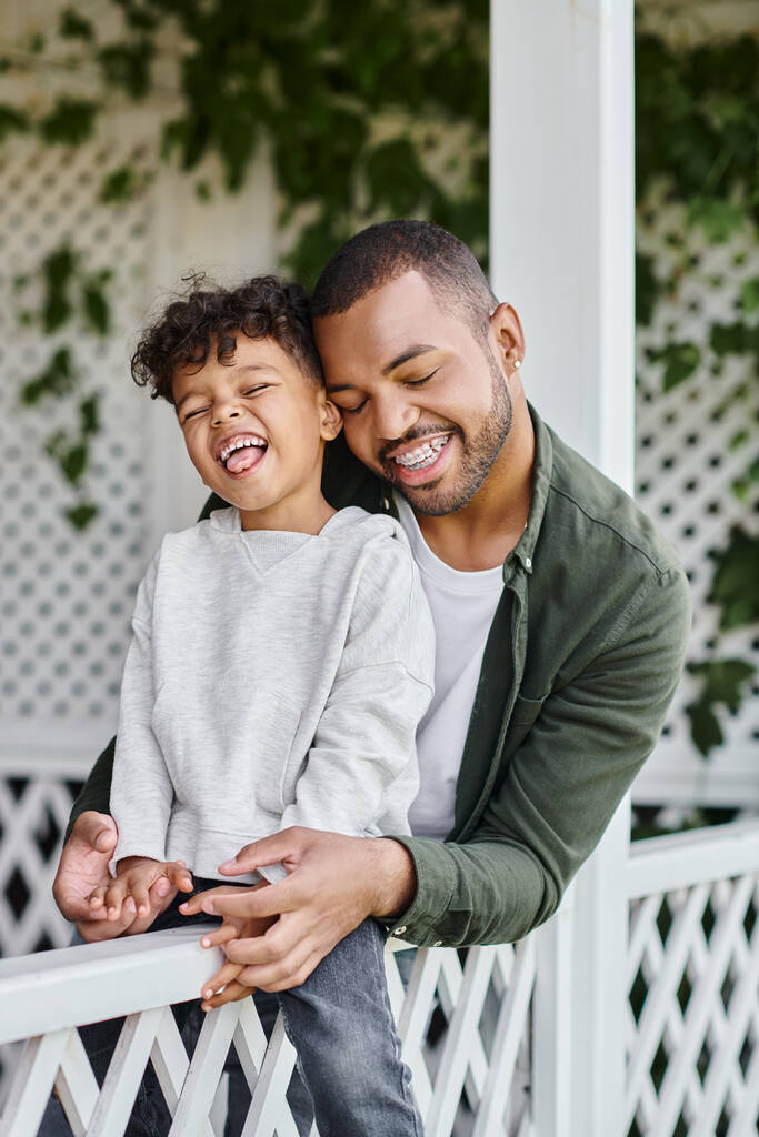 щасливий афроамериканський чоловік в брекетах обіймає кучерявого сина, сидячи на ганку з білим парканом - Фото, зображення