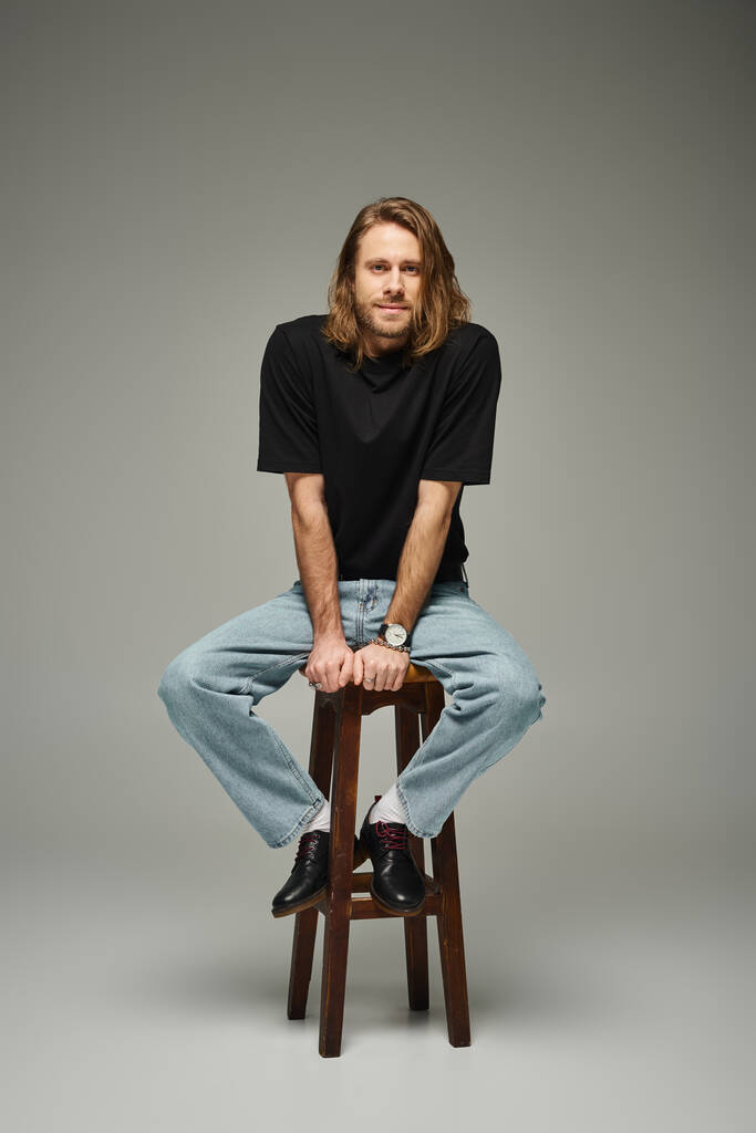 longitud completa del hombre barbudo de buen aspecto con el pelo largo sentado en jeans y camiseta en el taburete alto - Foto, imagen