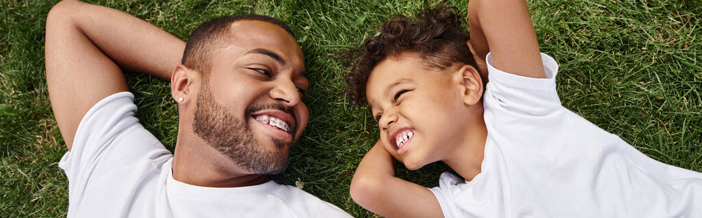 裏庭に緑の芝生に横たわる陽気なアフリカ系アメリカ人の父親と息子のトップビュー,家族の旗 - 写真・画像