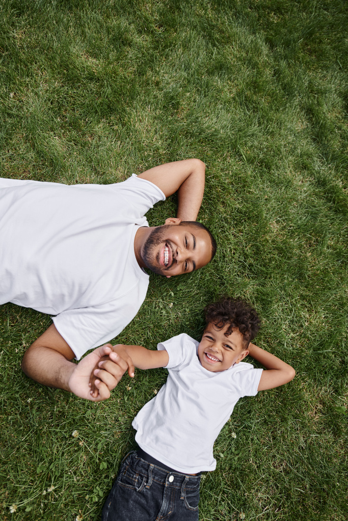 top view of pleased african american father and son lying on green lawn on backyard, family photo - Photo, Image