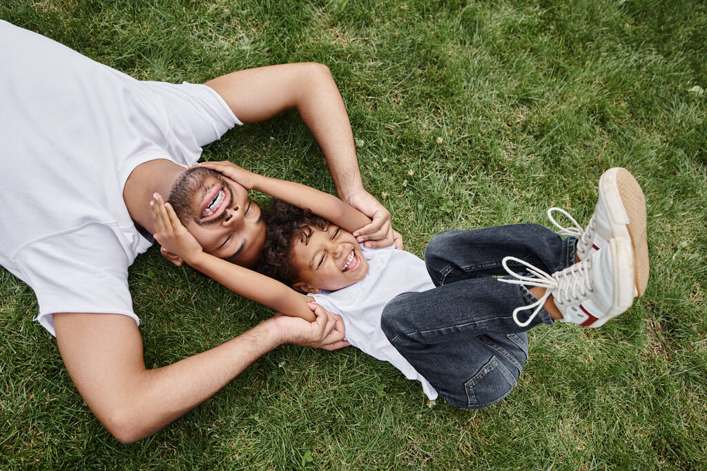 top view of cheerful african american father and son lying on green grass on backyard, family photo - Photo, Image
