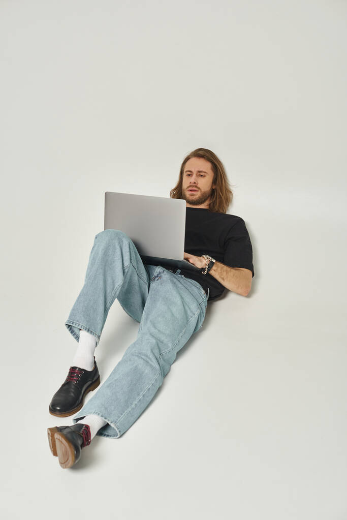 όμορφος άντρας με μακριά μαλλιά και γενειάδα ακουμπά στον λευκό τοίχο και χρησιμοποιεί φορητό υπολογιστή σε γκρι φόντο - Φωτογραφία, εικόνα