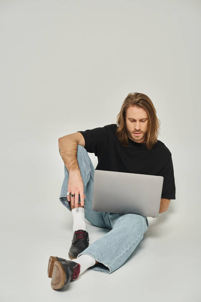 όμορφος ελεύθερος επαγγελματίας με μακριά μαλλιά και γενειάδα κάθεται στο πάτωμα και χρησιμοποιώντας φορητό υπολογιστή σε γκρι φόντο - Φωτογραφία, εικόνα