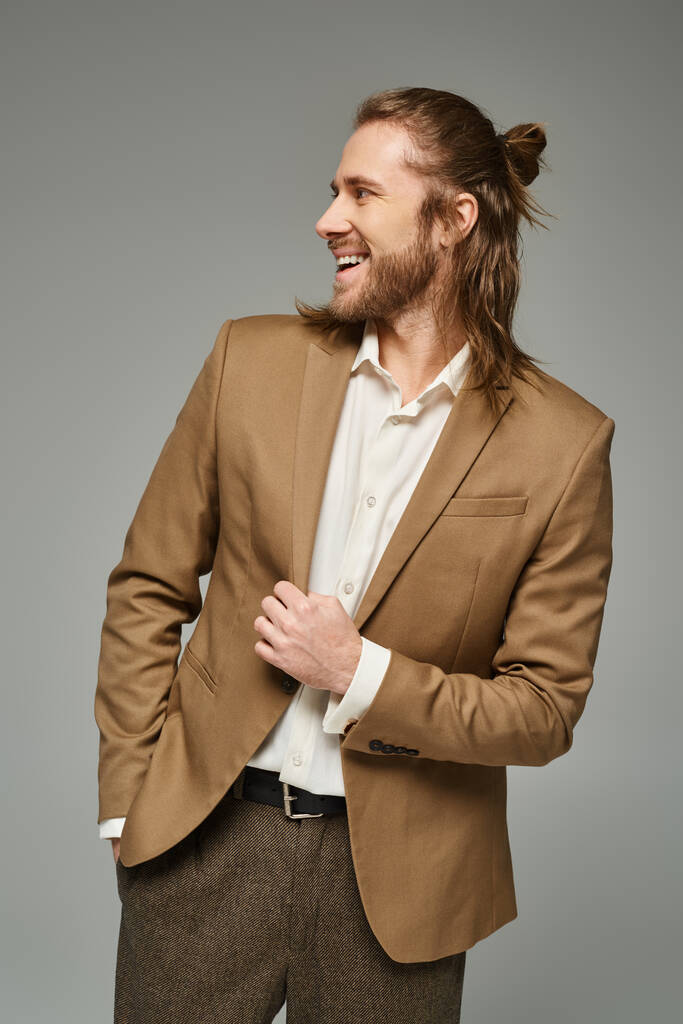 allegro uomo d'affari con barba e capelli lunghi in posa in blazer su sfondo grigio, abbigliamento formale - Foto, immagini