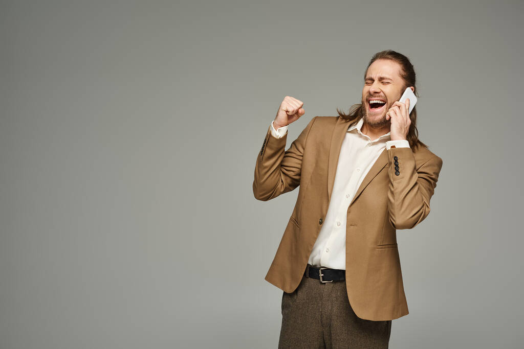 hombre de negocios barbudo guapo y emocionado en traje formal hablando en el teléfono inteligente sobre fondo gris - Foto, imagen