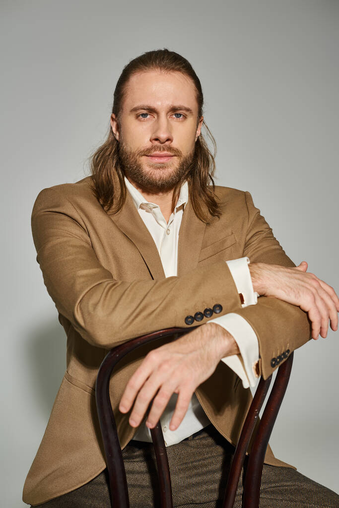 bonito barbudo empresário no bege formal traje sentado no madeira cadeira no cinza fundo - Foto, Imagem