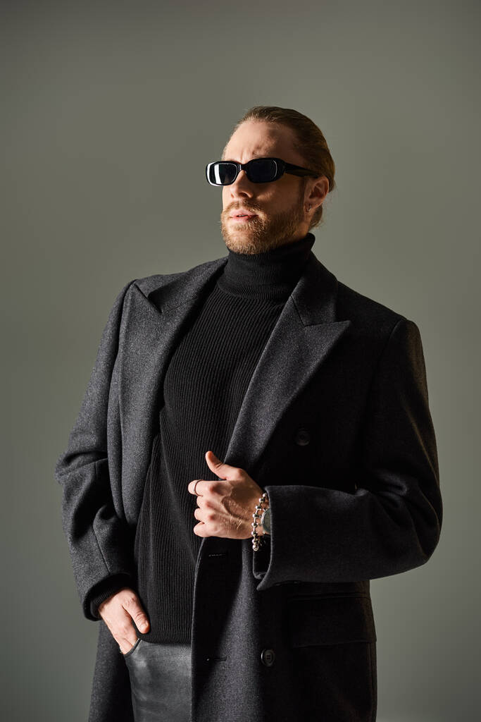 Porträt eines modischen Mannes mit trendiger Sonnenbrille und Mantel, der mit der Hand in der Tasche auf grau posiert - Foto, Bild