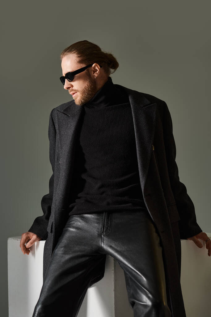 retrato de hombre guapo en gafas de sol de moda y abrigo de lana sentado en cubo blanco sobre fondo gris - Foto, imagen