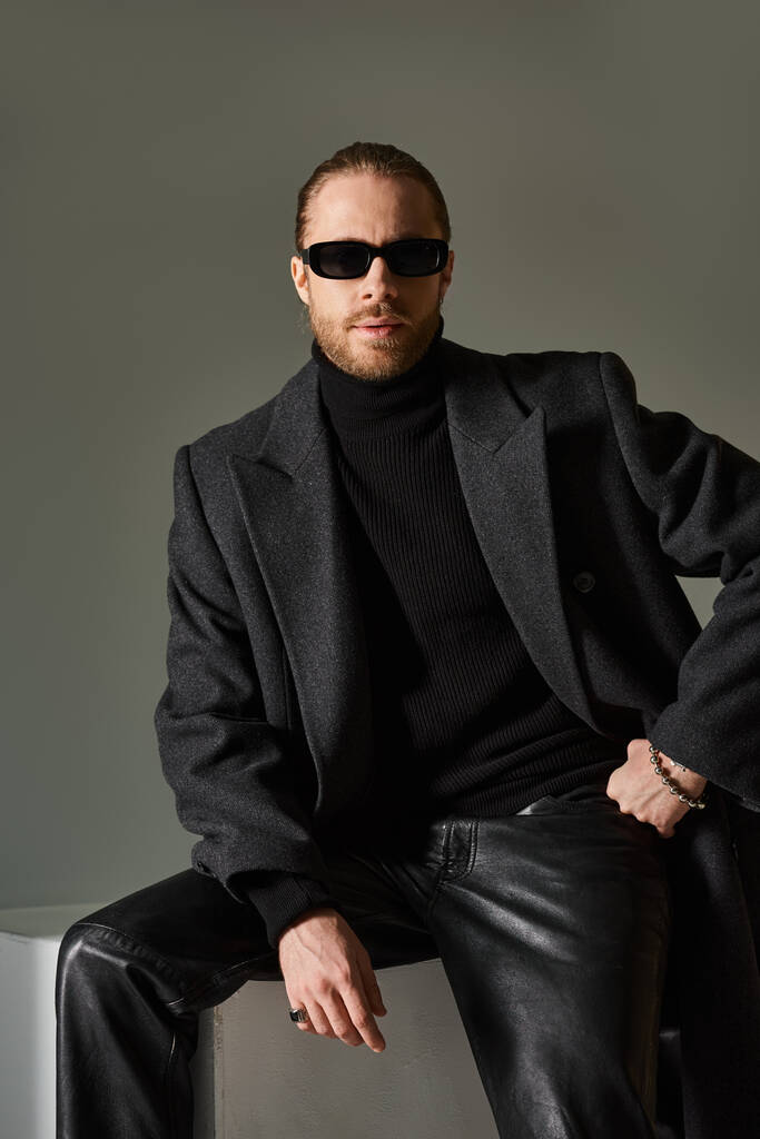 Porträt eines modischen Mannes mit trendiger Sonnenbrille und Mantel auf weißem Würfel vor grauem Hintergrund - Foto, Bild