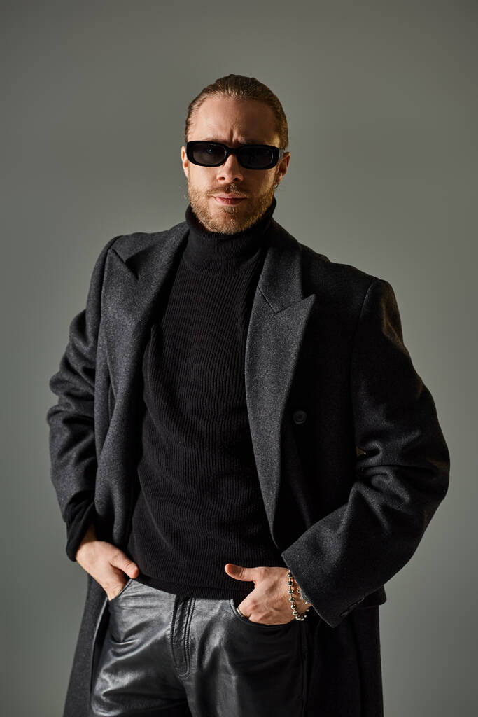 retrato de guapo modelo masculino en gafas de sol de moda y atuendo negro de pie con las manos en los bolsillos - Foto, Imagen