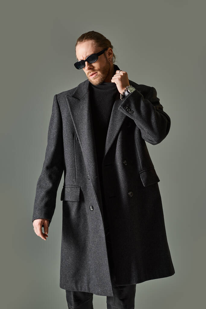 トレンディなサングラスと黒の服装でハンサムなひげ付き男 タートルネックの襟を調整 - 写真・画像