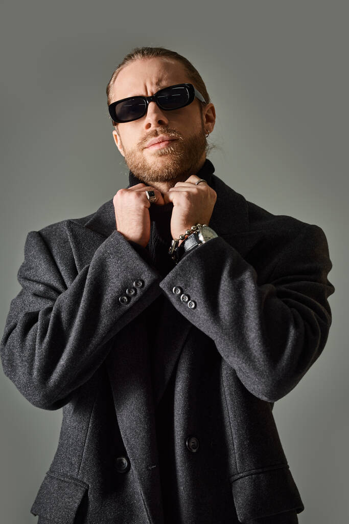pohledný vousatý muž v módních slunečních brýlích a černém oblečení upravující límec roláku - Fotografie, Obrázek