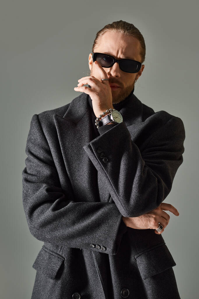 stylowy i brodaty mężczyzna w modnych ciemnych okularach przeciwsłonecznych i czarnym stroju pozowanie na szarym tle - Zdjęcie, obraz