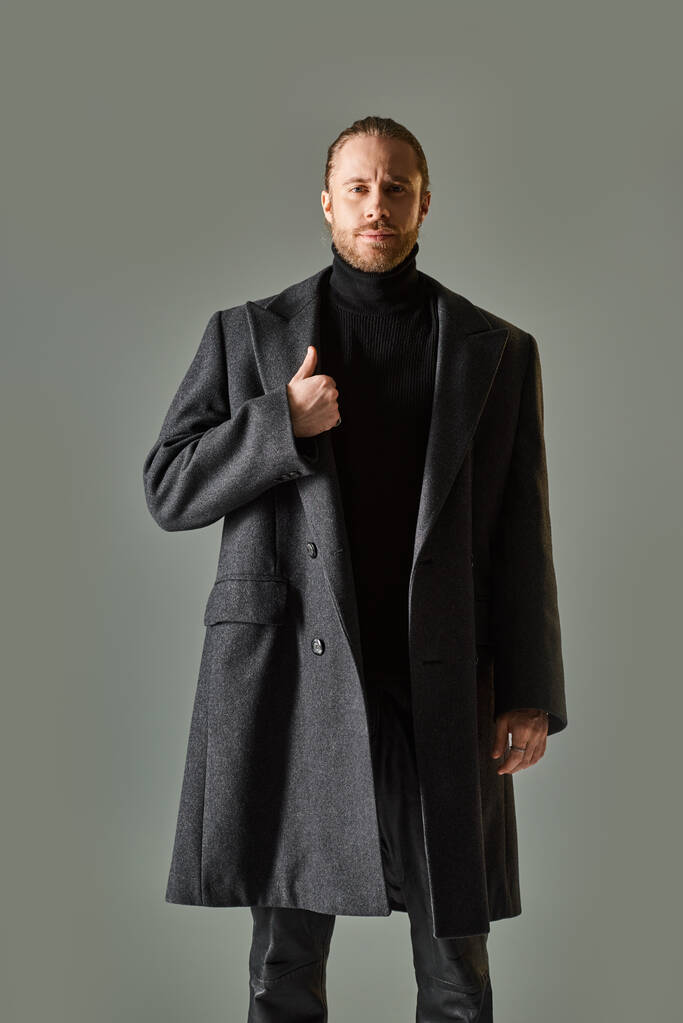 jóképű és szakállas férfi divatos fekete öltözet és gyapjú kabát pózol szürke háttér - Fotó, kép