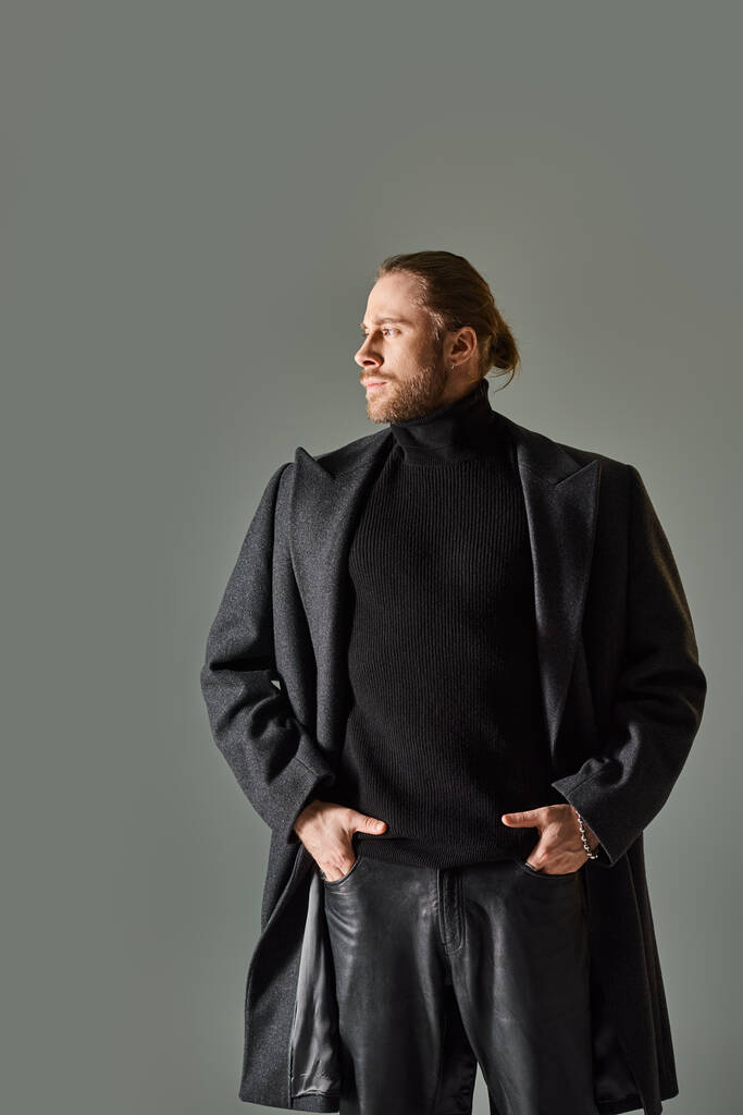 красивый и бородатый мужчина в модном черном одеянии и шерстяном пальто, смотрящий в сторону на сером фоне - Фото, изображение