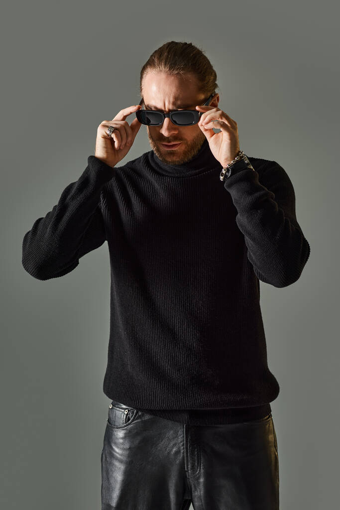 brodaty mężczyzna w modnych okularach przeciwsłonecznych, swetrze z golfem i czarnych skórzanych spodniach pozujących na szaro - Zdjęcie, obraz