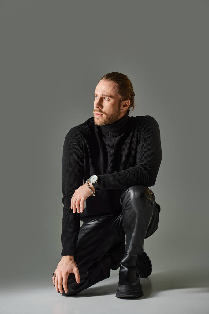 comprimento total do homem barbudo em camisola gola alta e calças de couro preto sentado no fundo cinza - Foto, Imagem