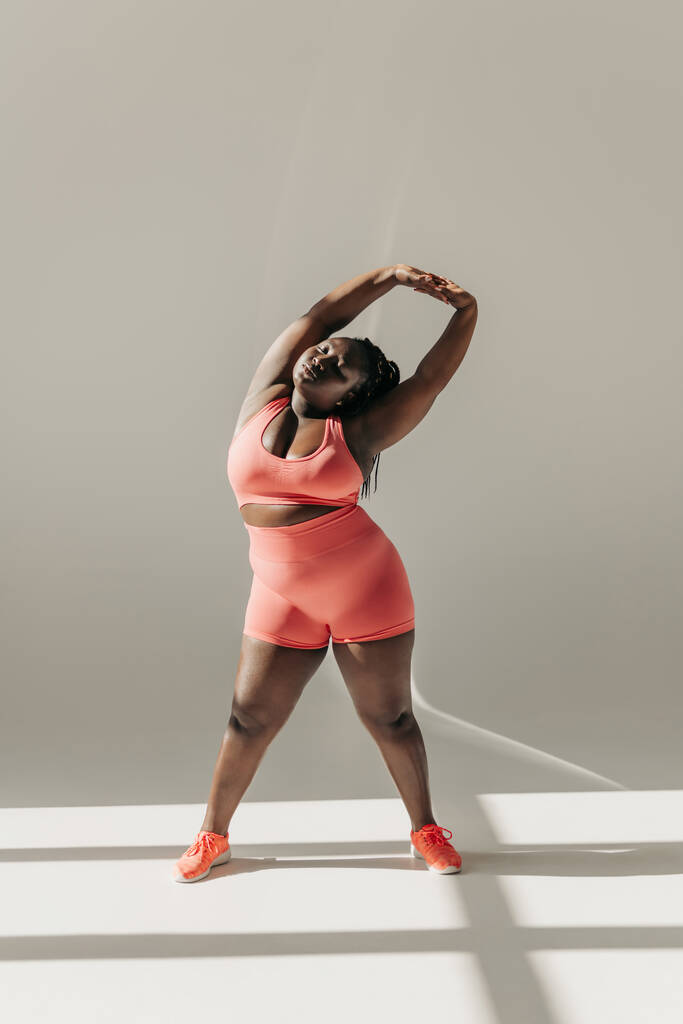 Pełna długość pewność siebie African plus size kobieta w odzieży sportowej robi ćwiczenia rozciągające w studio fitness - Zdjęcie, obraz