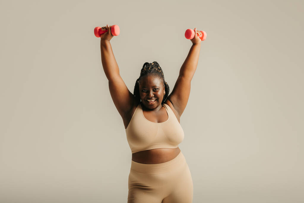 Spor kıyafetleri içinde kıvrımlı güzel bir Afrikalı kadın stüdyo arka planında egzersiz yaparken kendine güven yayıyor. - Fotoğraf, Görsel
