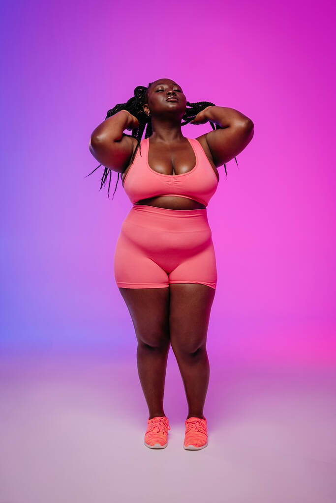 Longitud completa de mujer africana de tamaño grande en ropa deportiva ajustando dreadlocs contra fondo colorido - Foto, imagen