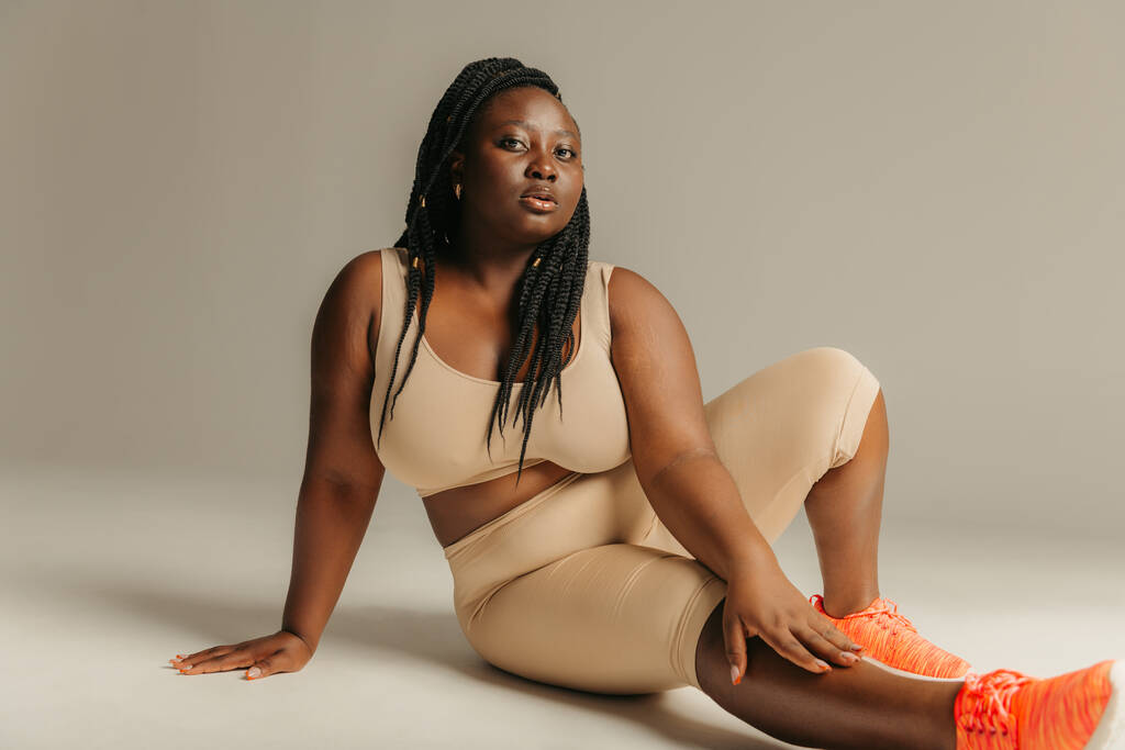 Молодая африканка в спортивной форме выглядит уверенно, сидя на студийном фоне - Фото, изображение