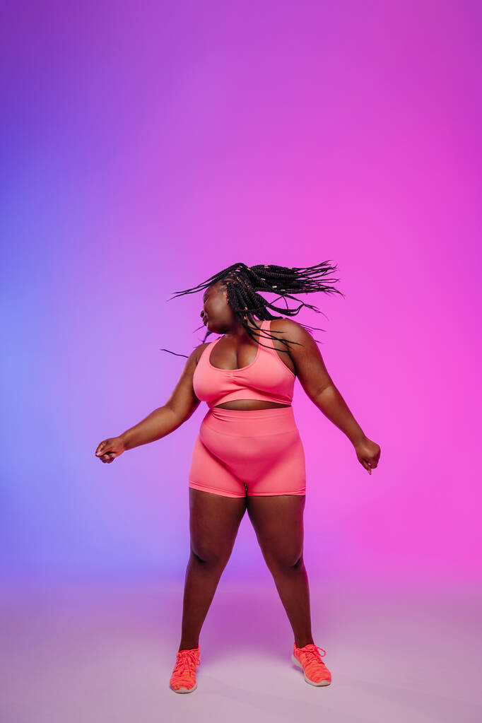 Schöne afrikanische Plus-Size-Frau in Sportbekleidung tanzt und schüttelt Dreadlocs vor buntem Hintergrund - Foto, Bild