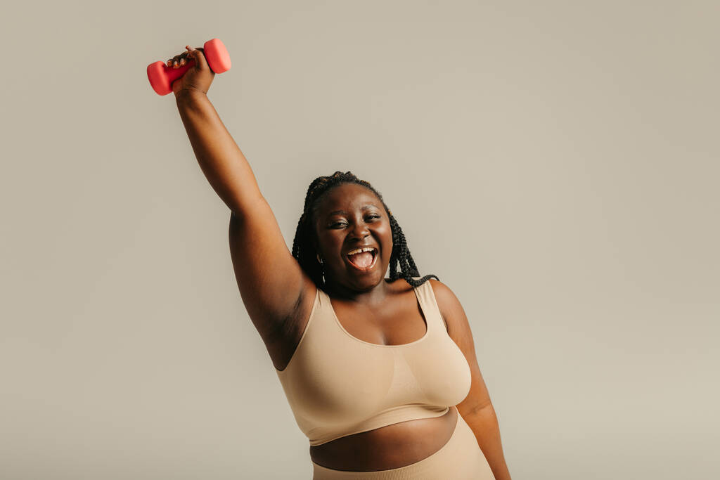Joyful plus size African woman in sportswear exercising with dumbbell on studio background - Photo, Image