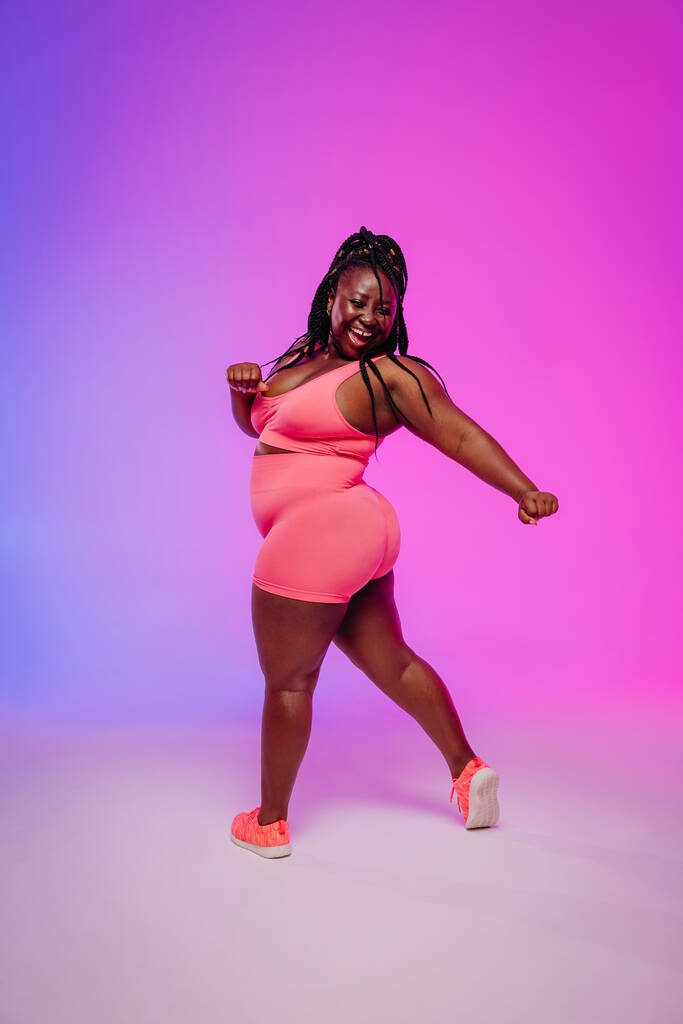 Joyful African plus size woman in sportswear dancing and radiating self-love against colorful background - Photo, Image