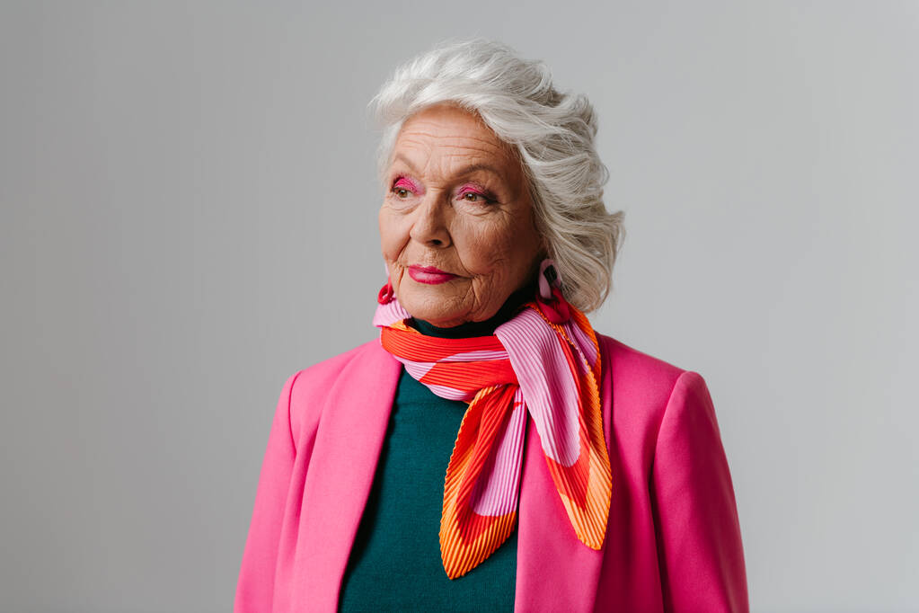Elegante oudere vrouw met make-up in modieuze kleding en stralende eigenliefde op grijze achtergrond - Foto, afbeelding