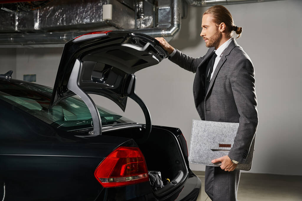 彼の車のトランクを閉じるポニーテールの洗練されたシックなスーツの魅力的なスタイリッシュなビジネスマン - 写真・画像