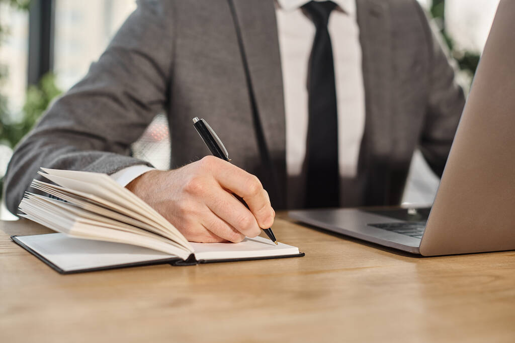 vista recortada del hombre de negocios en la escritura de ropa formal en el cuaderno mientras está sentado en el lugar de trabajo en la oficina - Foto, Imagen