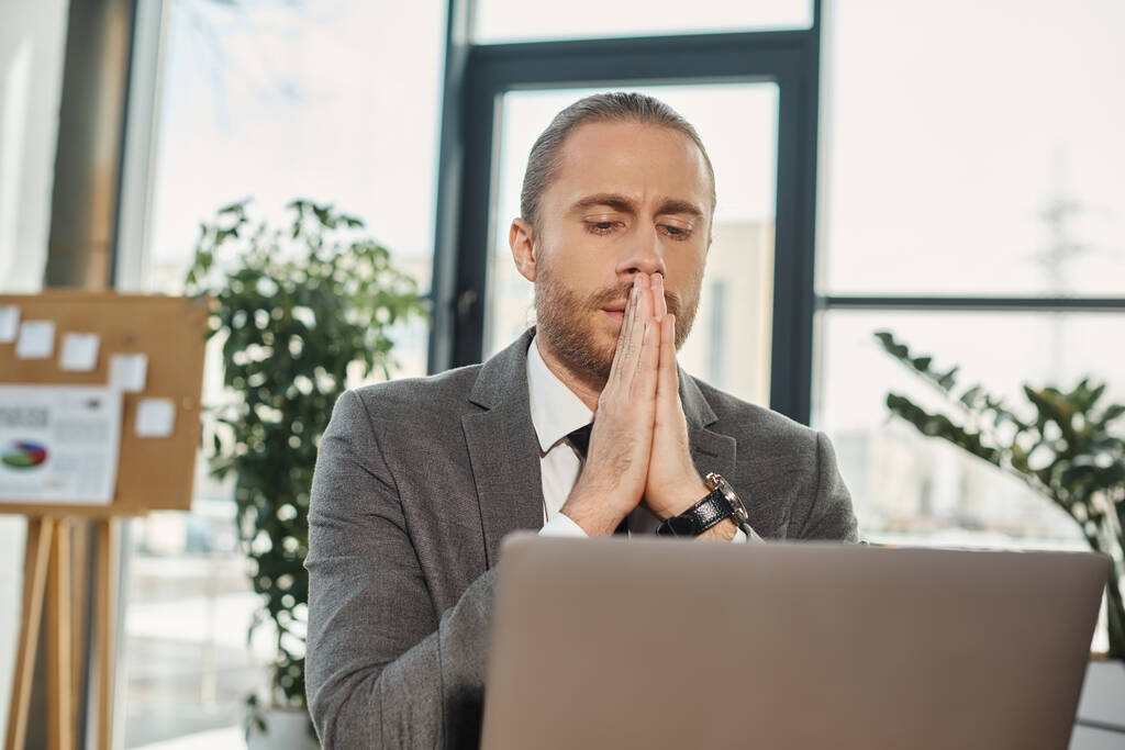 обеспокоенный бизнесмен сидит с молитвенными руками и смотрит на ноутбук во время работы в офисе - Фото, изображение