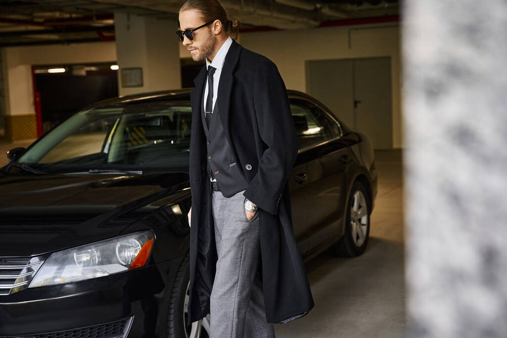 Attraktiver bärtiger Mann in schwarzem Mantel, der mit der Hand in der Tasche neben seinem Auto spaziert, Geschäftsidee - Foto, Bild