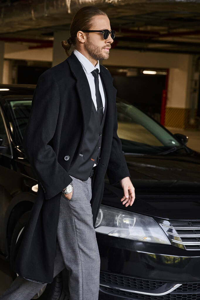 plano vertical de atractivo hombre de negocios en abrigo negro con gafas de sol caminando con la mano en el bolsillo - Foto, imagen
