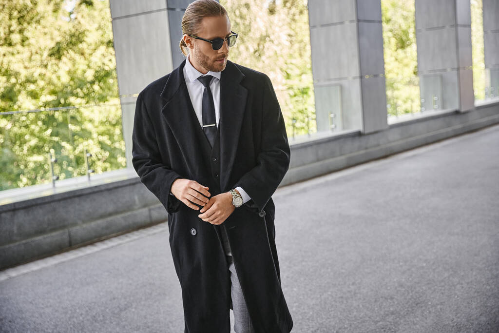 красивий бородатий чоловік в чорному елегантному пальто з сонцезахисними окулярами, позує на відкритому повітрі, бізнес-концепція - Фото, зображення