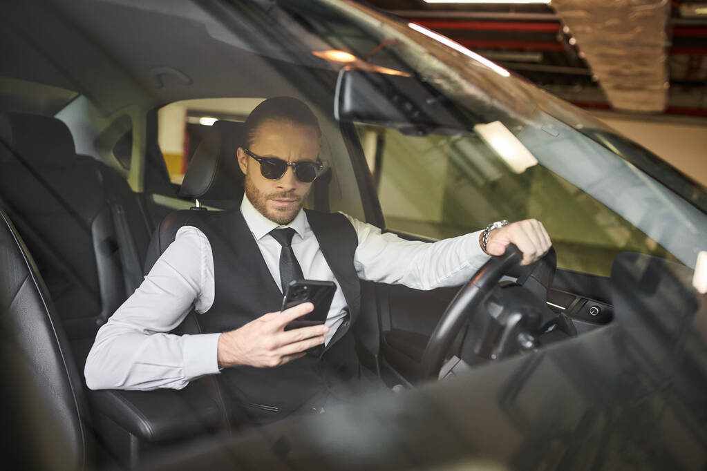 красивый очаровательный бизнесмен в черном жилете в солнечных очках, смотрящий на телефон за рулем - Фото, изображение