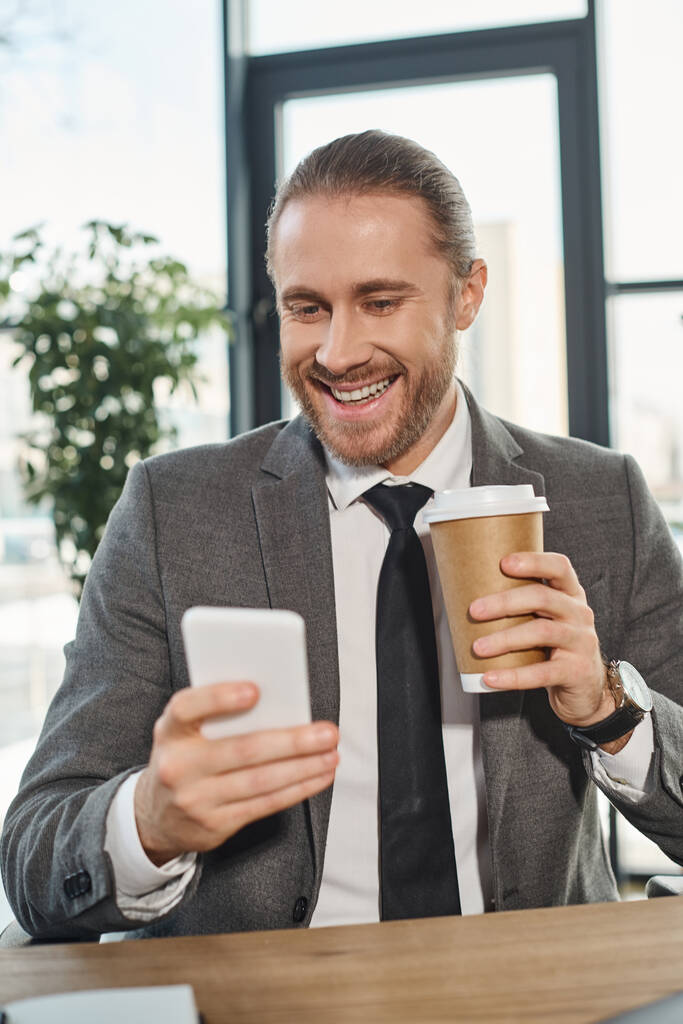 Χαρούμενος επιχειρηματίας με γκρι κοστούμι κρατώντας χάρτινο κύπελλο και δικτύωση σε smartphone στο γραφείο - Φωτογραφία, εικόνα