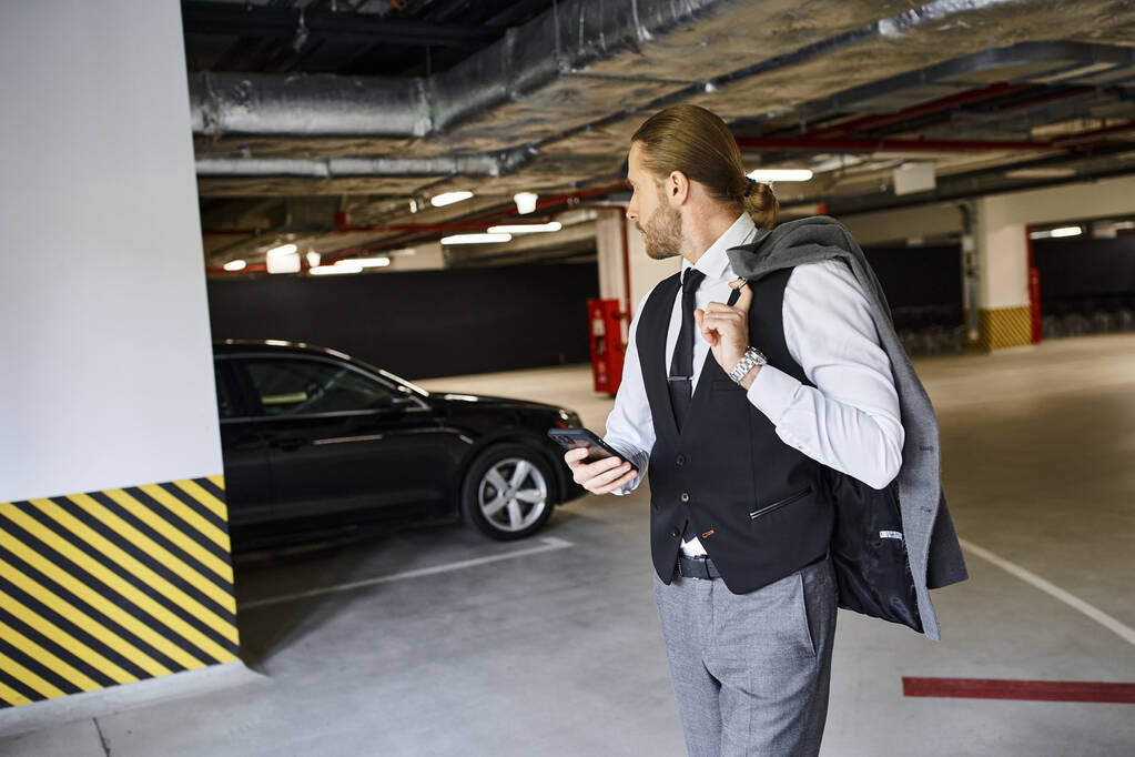 όμορφος γενειοφόρος άνδρας με μαύρο γιλέκο κρατώντας κινητό τηλέφωνο και κοιτάζοντας το αυτοκίνητό του, επιχειρηματική ιδέα - Φωτογραφία, εικόνα